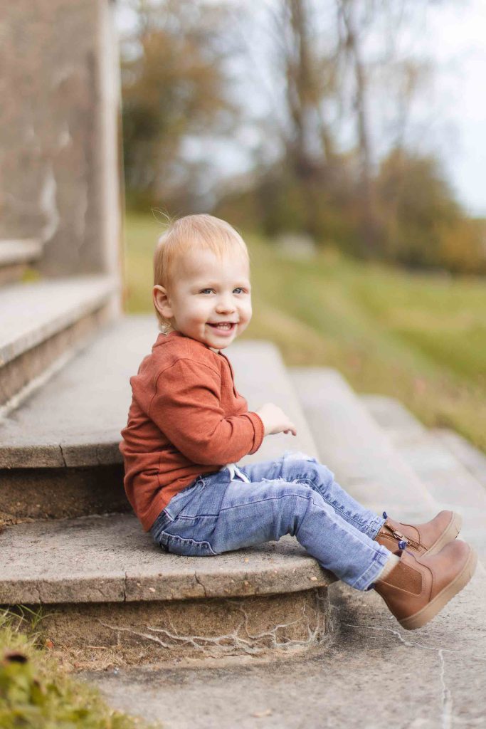 A young child wearing a red long-sleeve shirt, blue jeans, and brown ankle boots sits smiling on the outdoor stone steps of Belair Mansion with a blurred green field and trees in the background.