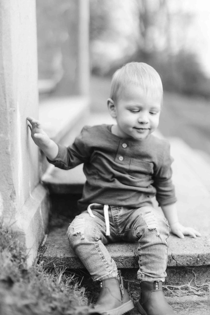 A black and white portrait captures a young child in Maryland sitting on outdoor steps. The child, donning a long-sleeve shirt, ripped jeans, and boots, looks down with one hand resting on the step—a perfect moment from a mini-session.