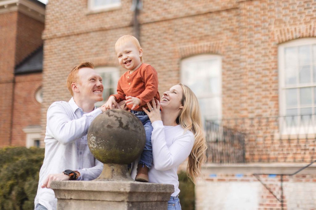 A man and a woman hold up a smiling toddler standing on a stone pedestal in front of a brick building during a mini-session in Bowie, Maryland.