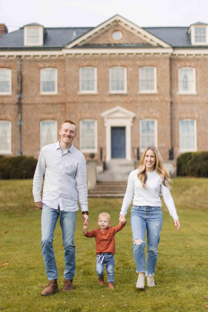 A family walks hand in hand with a young child between them on a lawn in front of a large brick building, capturing the perfect moment during their Maryland mini-session.