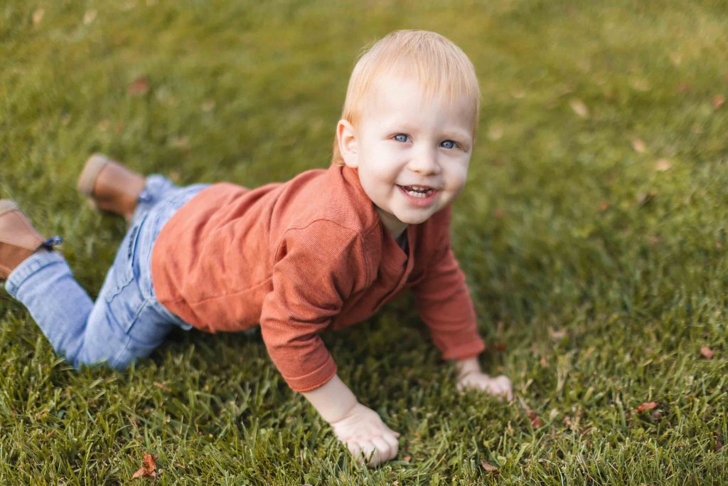 A young child wearing a red long-sleeve shirt and blue jeans is crawling on the grass, looking at the camera and smiling, at a family picnic near the historic Belair Mansion in Maryland.