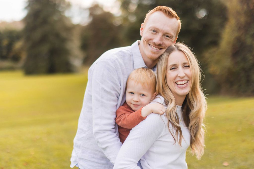 A smiling man, woman, and young child stand together in an outdoor setting at what appears to be a mini-session. The child is between the adults, being held by the woman. Trees and grass are visible in the background, capturing a serene moment perhaps near Belair Mansion in Maryland.