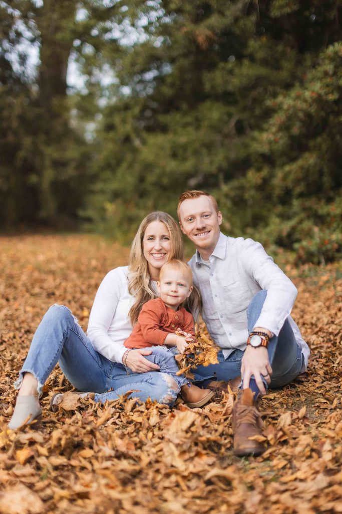 A family of three sits on the ground covered with autumn leaves, smiling at the camera. The parents are dressed in casual white tops, and the child is wearing a rust-colored shirt. Trees are in the background, creating a perfect portrait reminiscent of a scene near Belair Mansion in Bowie.
