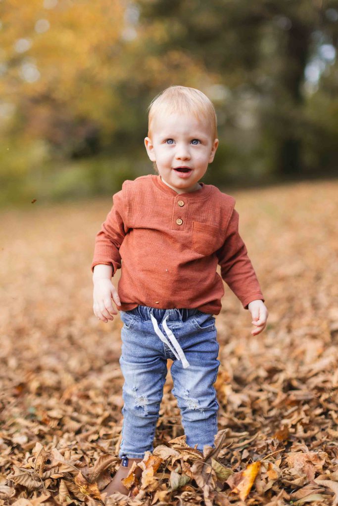 A toddler with blonde hair stands in a field of fallen leaves, wearing a rust-colored long-sleeve shirt and blue jeans. The scene evokes the charm of a Bowie portrait with trees displaying autumnal foliage in the blurred background.