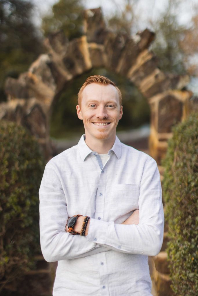A red-haired man wearing a white shirt stands with folded arms in front of a rustic stone archway at Belair Mansion in Bowie, smiling. Bushes flank the archway, adding a charming touch to the historic family estate.