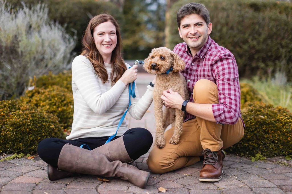 A man and a woman are outdoors in Maryland, both smiling while holding a small dog. The woman is sitting on the ground holding a leash, and the man is crouching down next to the dog, capturing the perfect moment during their mini-session.