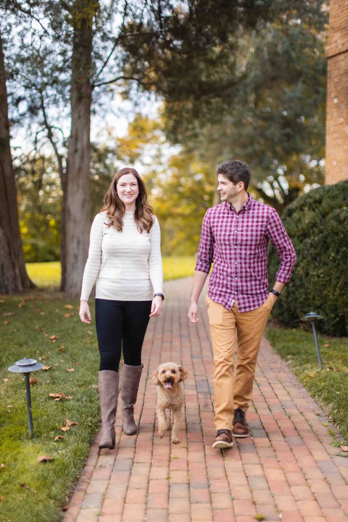 A man and woman walk down a brick pathway in Maryland with a small dog between them. Trees and bushes are in the background. They are smiling, casually dressed, and enjoying their mini-session together.