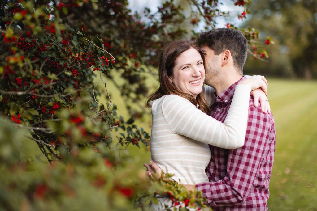 A couple is embracing outdoors near a berry-laden bush. The woman is smiling while the man, dressed in a red checkered shirt, has his face partially hidden. This tender moment was captured during a mini-session in Bowie, preserving a beautiful memory for their family.