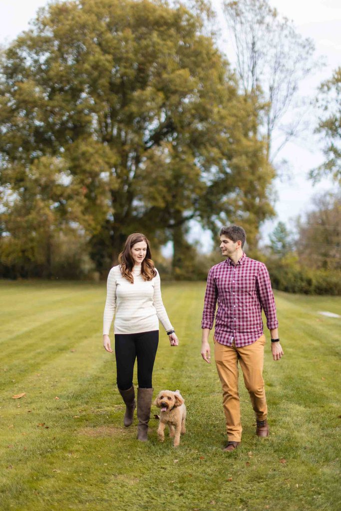 A man and woman walk a small dog on a grassy path with trees in the background, capturing a serene moment perfect for a mini-session at Bowie’s historic Belair Mansion.