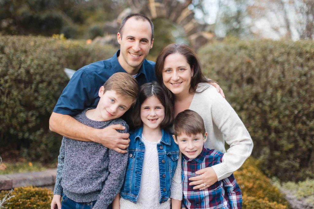 A group photo of a family during a mini-session in Maryland, featuring two adults and three children standing close together and smiling at the camera, with greenery in the background.