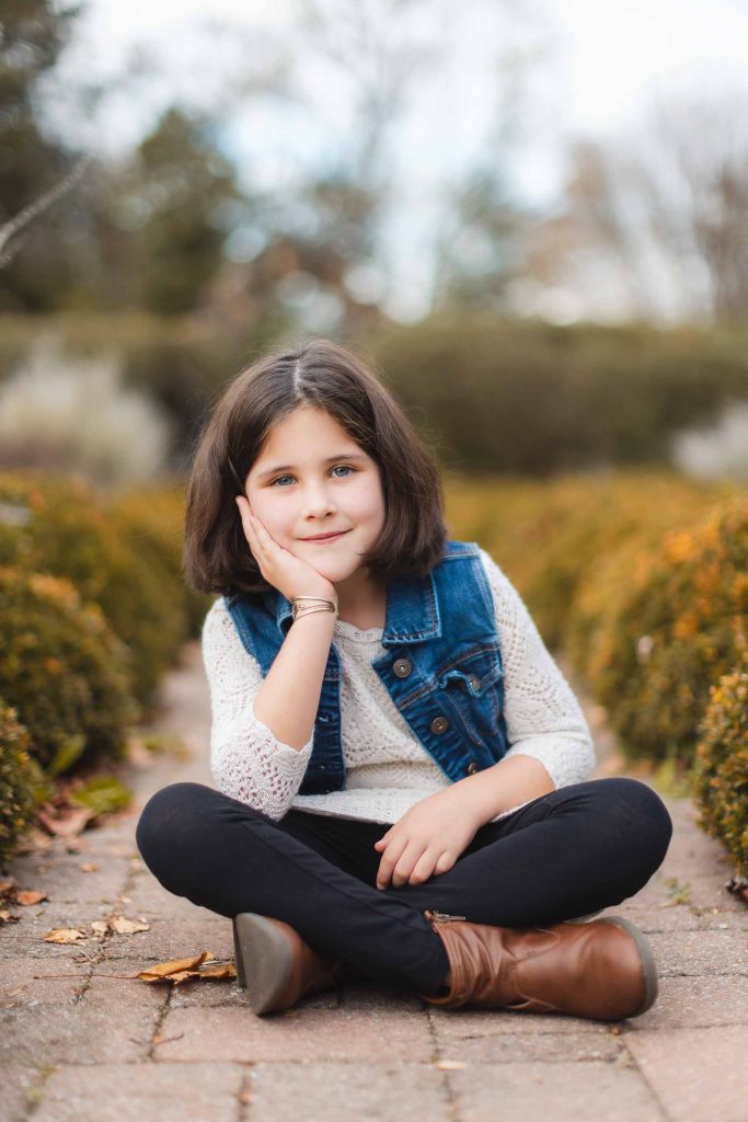 A girl with dark hair sits cross-legged on a paved path, resting her head on her hand. She is wearing a denim vest over a white sweater, reminiscent of Bowie's relaxed style, and there are hedges in the background. It's the perfect scene for a portrait mini-session.