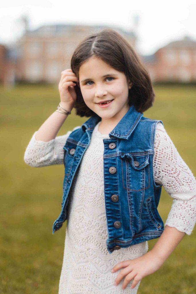 Young girl with dark hair, wearing a white dress and denim vest, stands outdoors on a grassy field near Belair Mansion, smiling slightly and touching her hair. Blurred buildings are visible in the background of the mini-session portrait.