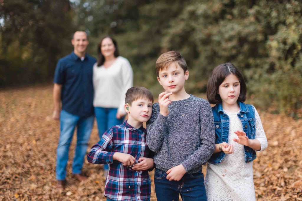 Three children stand together in a wooded area with fallen leaves during a family mini-session, while two adults stand blurred in the background.