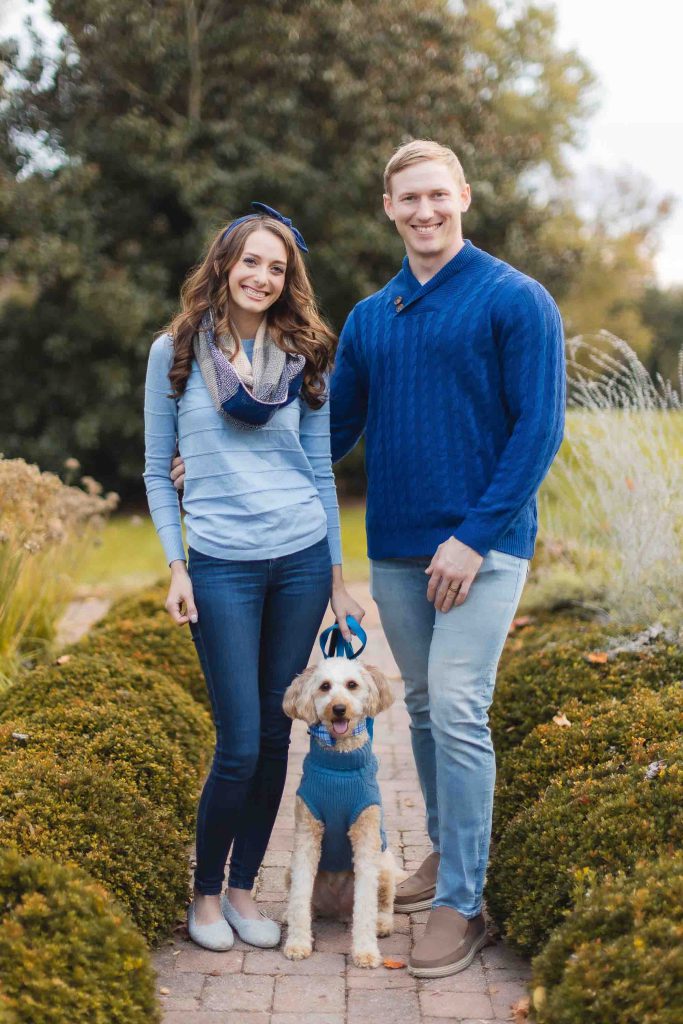 A woman and a man, both wearing blue tops, stand on a garden path at Belair Mansion with their dog. The dog is also dressed in a blue sweater. Shrubs and trees are in the background, setting the perfect scene for a mini-session family photo.