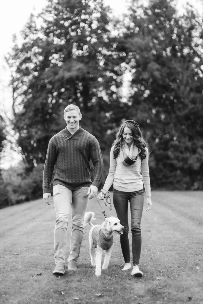 A man and a woman walk a dog on a leash in a grassy area with tall trees in the background. The image, capturing this serene family mini-session, is rendered in black and white.