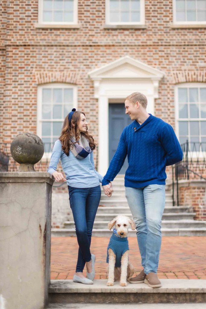 A family stands holding hands on the steps of a brick house, with a dog in a blue sweater sitting between them, capturing the perfect moment for their Maryland mini-session.