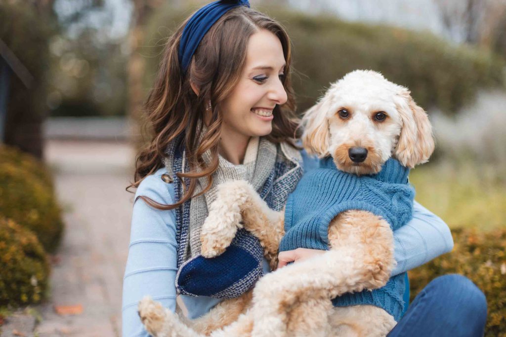A woman in a blue sweater and headband is sitting outdoors, holding a cream-colored dog wearing a matching blue sweater. Both seem content during their mini-session, with the blurred greenery of Belair Mansion in the background.