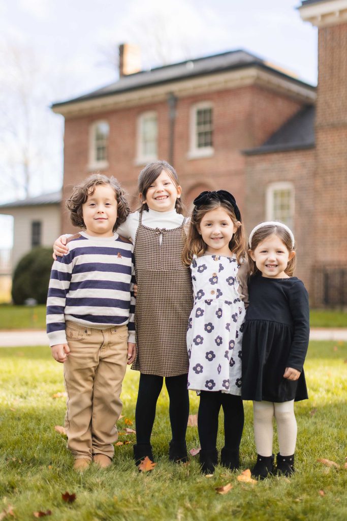 Four children stand side by side on the grass in front of Belair Mansion, smiling at the camera. The scene resembles a timeless family portrait with two girls in dresses and the other two children in long-sleeve tops and pants.