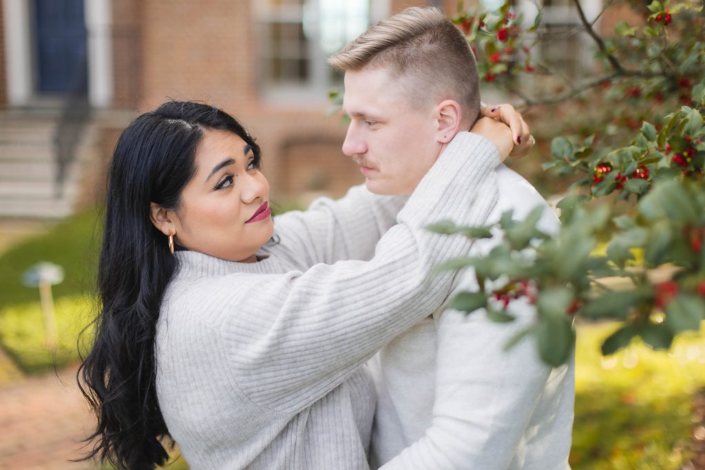 A woman and man, both wearing light-colored sweaters, stand close together outdoors at Belair Mansion, with the woman looking at the man and holding him around his neck. Red berries and green leaves are visible nearby, creating a picturesque portrait.