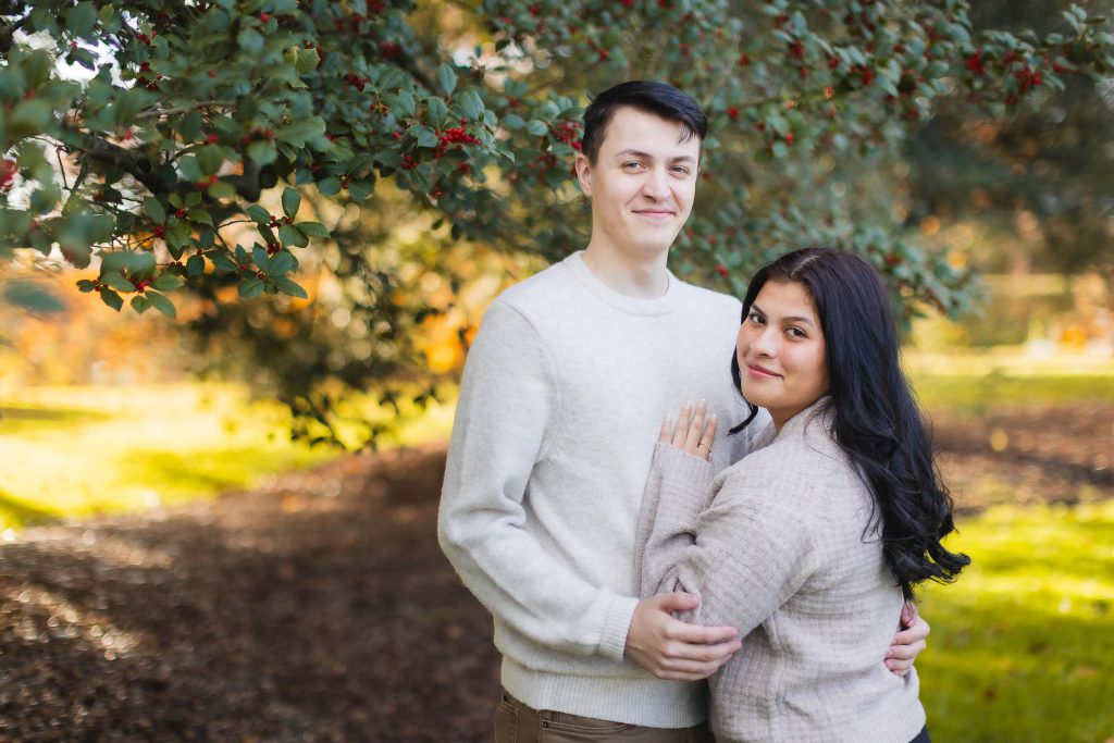 A man and a woman stand outdoors under a leafy tree, both wearing light-colored sweaters. The woman leans against the man as they look at the camera, creating a family portrait reminiscent of serene moments one might capture at Belair Mansion.