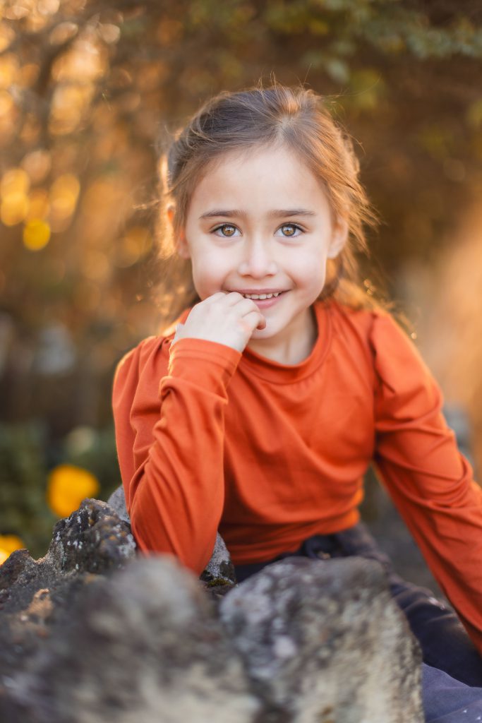 A child with brown hair wearing an orange long-sleeve shirt sits outdoors, smiling with one hand near their chin. The background shows blurred green foliage, reminiscent of a charming portrait that could grace the walls of Belair Mansion.