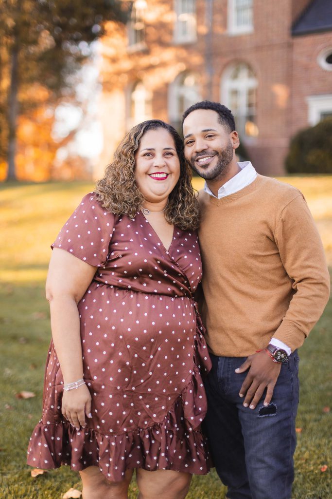 A woman in a brown polka-dot dress and a man in a tan sweater stand smiling together outdoors with the historic Belair Mansion and fall foliage in the background.