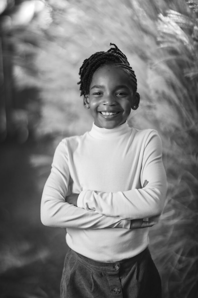 Black and white family portrait of a young person with a braided hairstyle, wearing a long-sleeve shirt, standing with arms crossed and smiling at the camera.