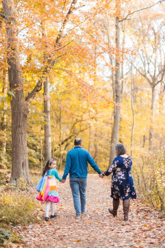 A family of three holding hands, walking down a forest path lined with autumn-colored trees in Greenspring Garden, Alexandria. The child is dressed in a vibrant, colorful outfit.