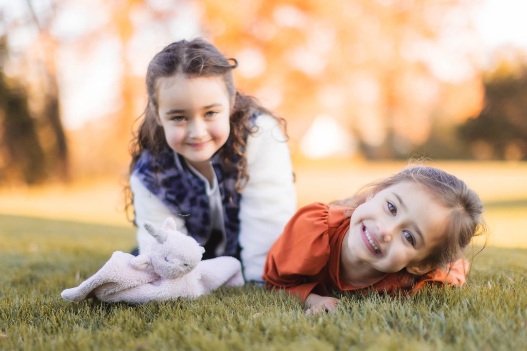 Two young girls lie on the grass with a plush toy, smiling at the camera, creating a heartwarming scene. The background is filled with blurry, autumn-colored trees near Belair Mansion, making it a perfect setting for family portraits and extended family gatherings.