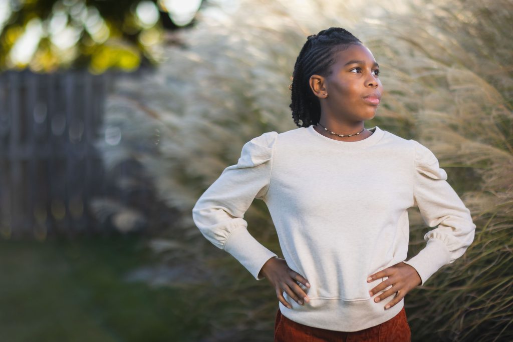 A person stands outside with hands on hips, wearing a light-colored sweatshirt and looking to the side. Tall grass and a wooden fence are in the background, creating a serene setting perfect for a family portrait.