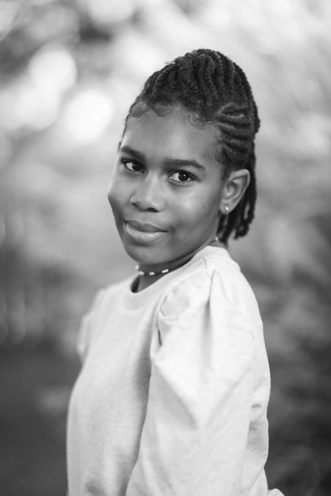 Black and white portrait of a young girl with braided hair, wearing a long-sleeved shirt, smiling softly at the camera, with a blurred outdoor background. The image captures a tender family moment.