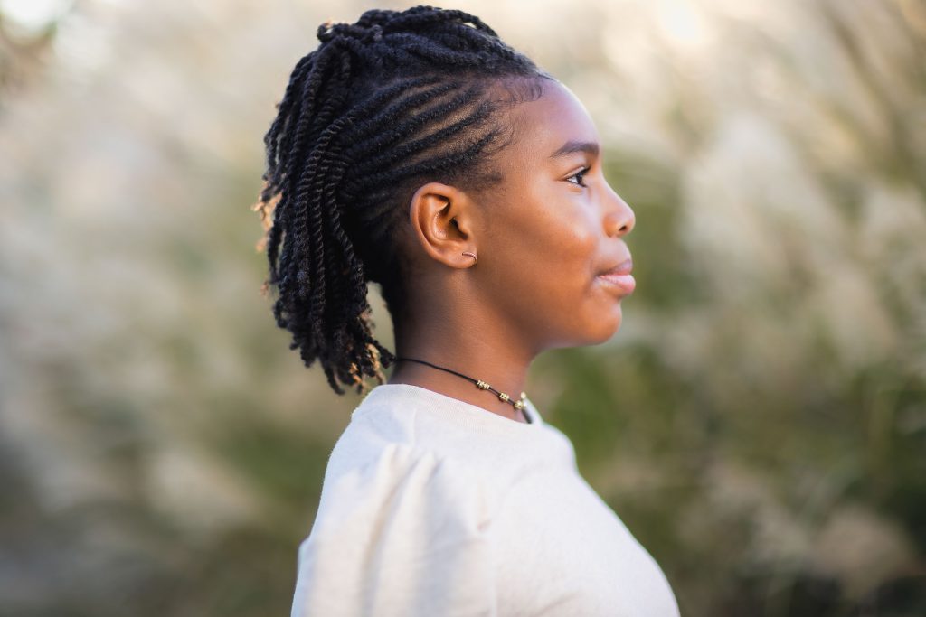 A person with braided hair stands outside, facing sideways. They are wearing a light-colored top and a necklace, creating an intimate portrait that captures the essence of family moments.