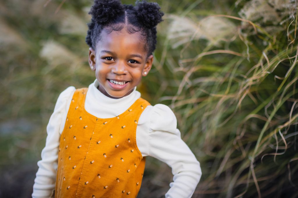 A young girl with puffball-style buns smiles at the camera in this charming family portrait, wearing a yellow dress over a white long-sleeve shirt, standing outdoors with greenery in the background.