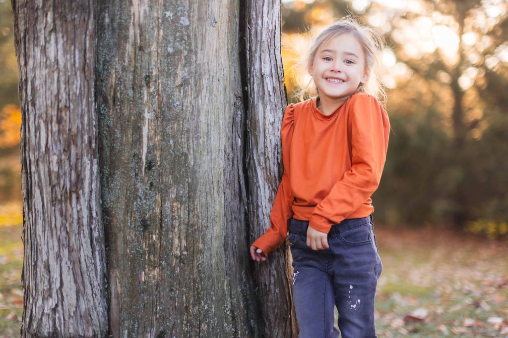 A young girl with a smile stands next to a large tree wearing an orange long-sleeve shirt and dark pants. The background features an outdoor setting with sunlight filtering through the trees, perfect for capturing timeless portraits reminiscent of family gatherings at Belair Mansion.