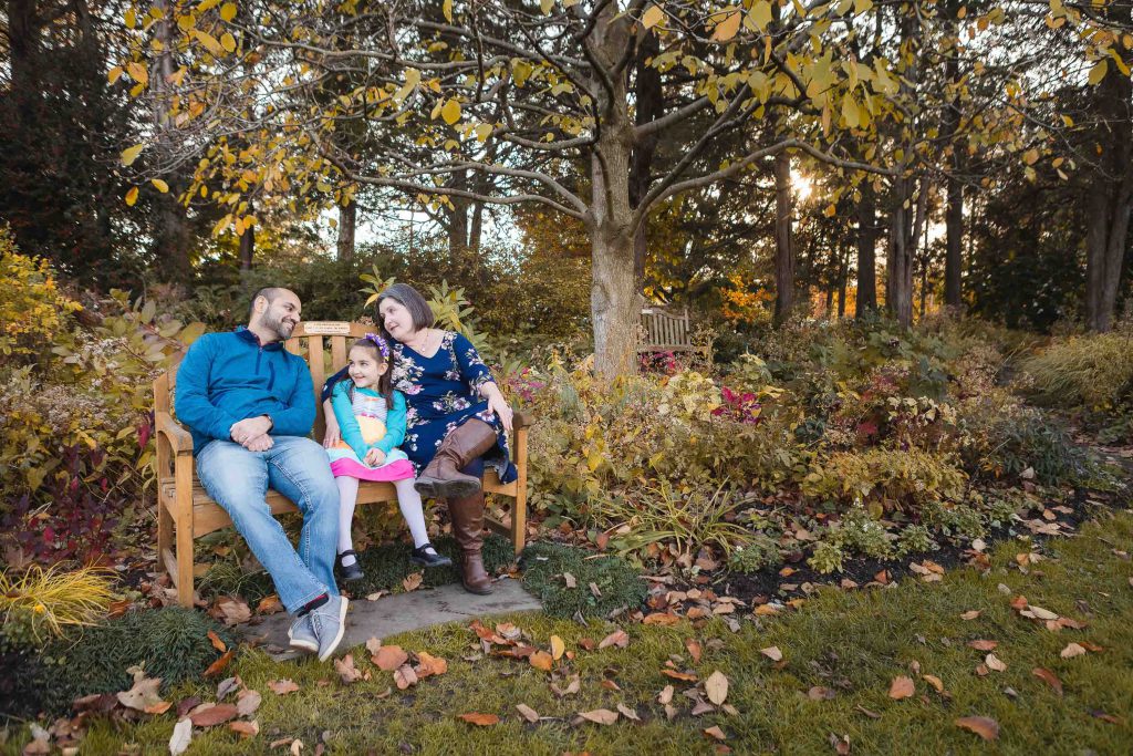 A man, woman, and young girl sit on a wooden bench in Virginia's Greenspring Garden with autumn foliage, surrounded by trees and shrubs. The adults look at the girl, who smiles while sitting between them in this lovely portrait.