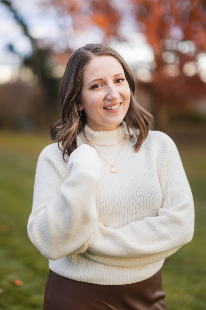 A portrait of a woman with shoulder-length brown hair wearing a white sweater and brown skirt smiles outdoors with autumn foliage in the background, creating a timeless memory at Belair Mansion.
