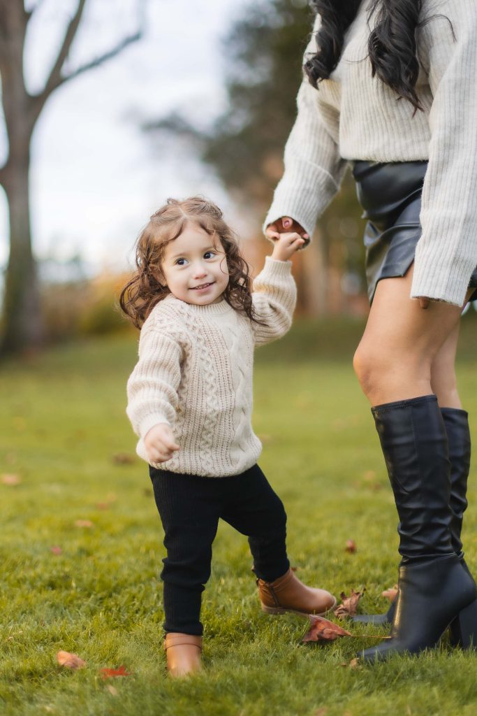 A young child with curly hair, wearing a beige sweater, holds an adult's hand and smiles while standing on the grass outdoors. The portrait captures the family moment perfectly as the adult, dressed in a cream sweater and black skirt with knee-high boots, enjoys a day out at Belair Mansion.