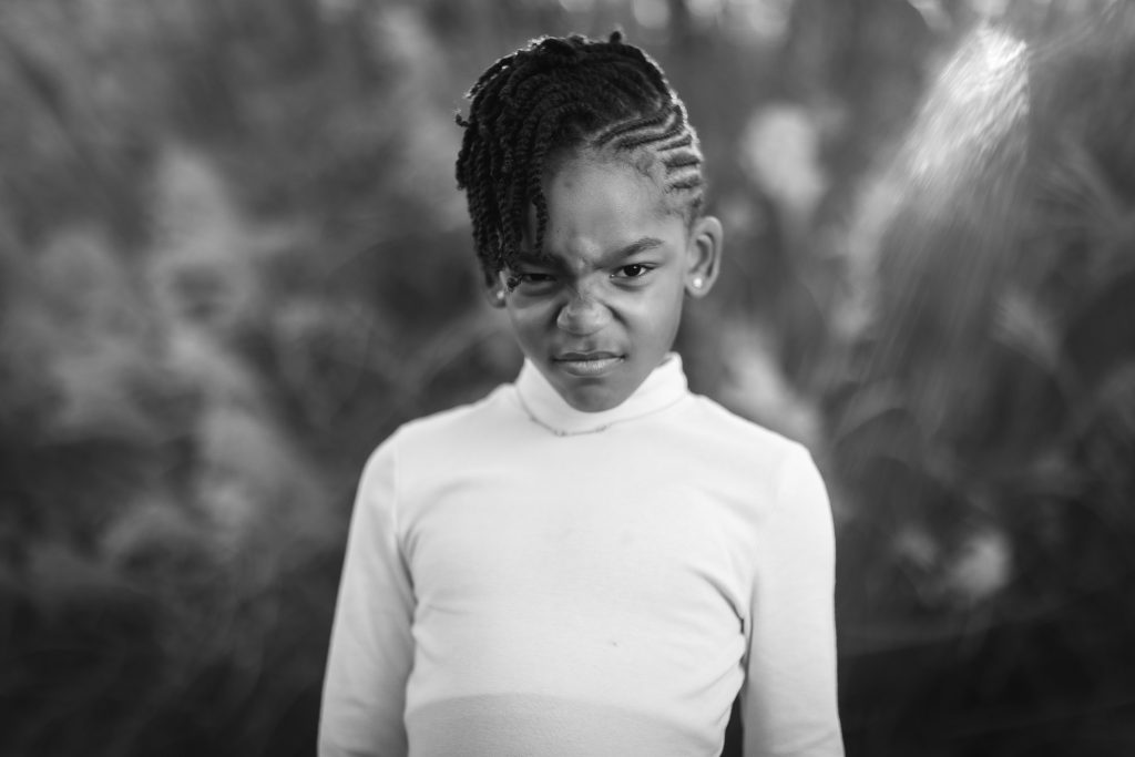 Black and white family portrait of a young girl with braided hair and a side-swept hairstyle, wearing a long-sleeve top, making a scrunched-up facial expression, and standing against a blurred natural background.