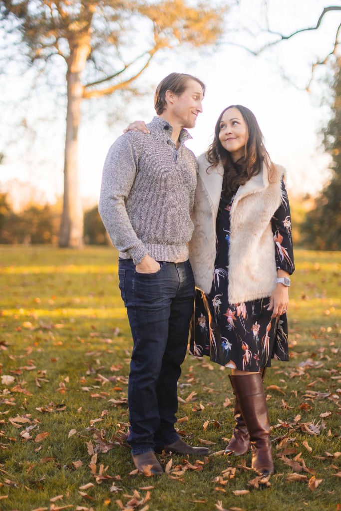 A man and woman stand on a grassy, leaf-strewn area with trees in the background, smiling and looking at each other. The man wears a grey sweater and jeans, while the woman is in a dress, vest, and boots. This could be an ideal spot for extended family portraits at Belair Mansion.