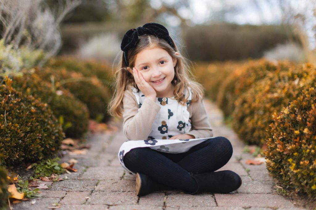A smiling girl with a hand on her cheek sits cross-legged on a pathway bordered by neatly trimmed bushes at Belair Mansion. She is wearing a floral dress, black tights, and a black headband. This portrait captures the serene beauty of family moments in an elegant setting.