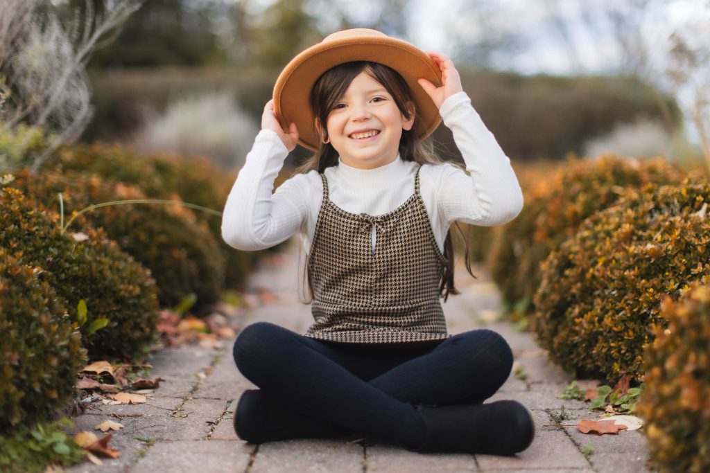 A young girl sits cross-legged on a brick pathway in front of Belair Mansion, smiling and adjusting a tan hat with both hands. She is wearing a white long-sleeve shirt, patterned dress, and black leggings. This charming portrait captures her joy as the surrounding bushes blur into the background.