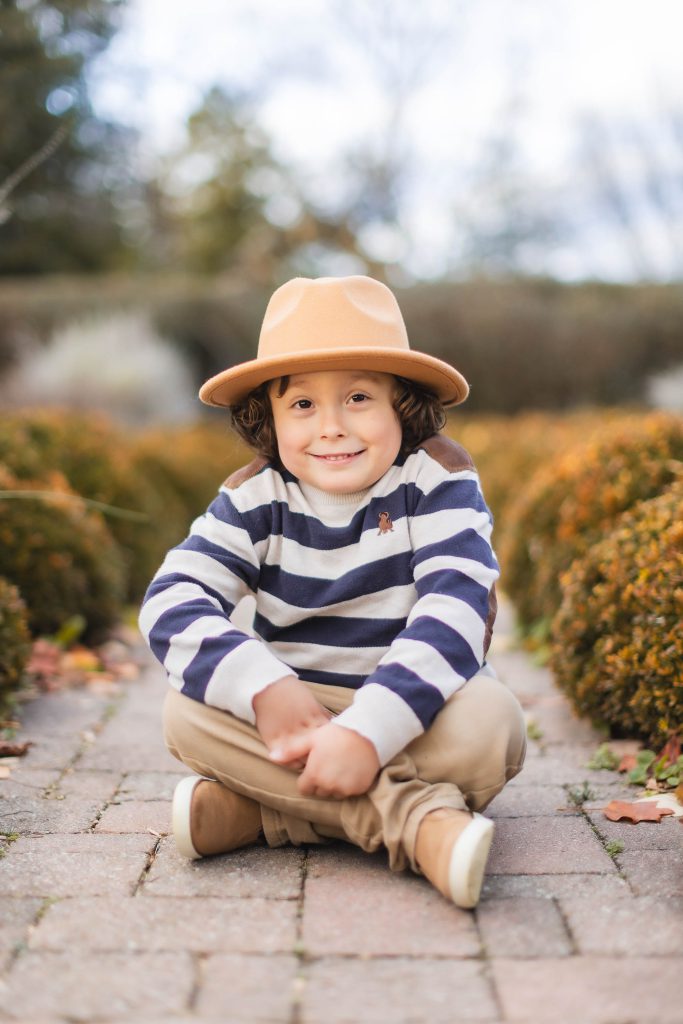 A child with curly hair sits cross-legged on a paved path, wearing a striped sweater, tan pants, tan shoes, and a tan hat. The background features blurred foliage and hedges, making it look like a serene portrait taken at Belair Mansion.