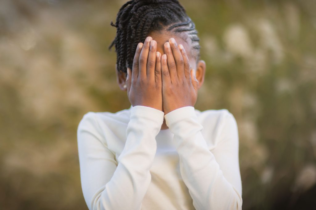 A child stands outdoors covering their face with both hands, wearing a white long-sleeve shirt, capturing a candid family moment.