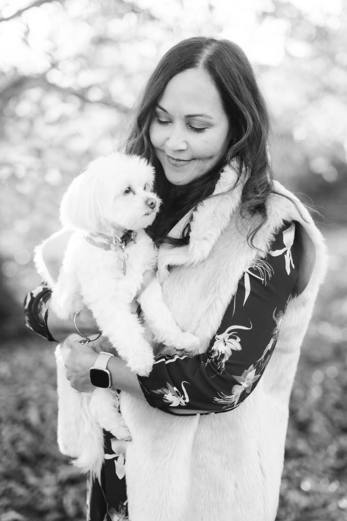 A woman wearing a fur-trimmed vest holds a small white dog while smiling outdoors in a black and white photo, capturing the essence of elegant portraits often seen at the Belair Mansion.