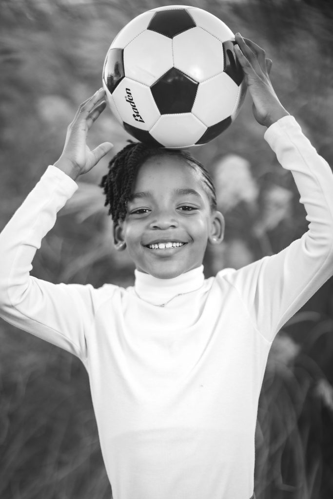 A black and white portrait of a child smiling and holding a soccer ball above their head with both hands. The child, wearing a long-sleeve shirt, radiates joy, capturing the essence of family fun.