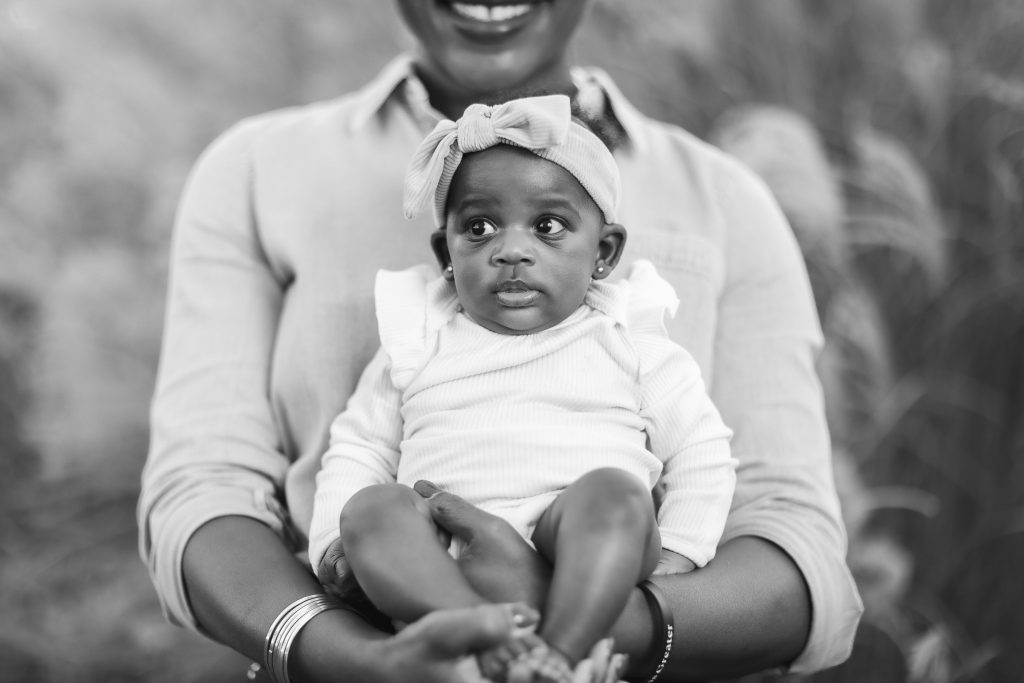A timeless black and white portrait captures a smiling person holding a baby dressed in a light-colored outfit with a headband, embodying the essence of family. The person's face is partially visible, adding an intimate touch to the cherished moment.