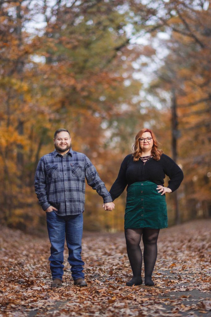 A man and a woman stand holding hands on a leaf-covered path in a forest during autumn. The woman has red hair and wears a black top and green skirt, while the man wears a plaid shirt and jeans. The moody dusk light casts an eerie glow over the scene, adding a touch of mystery to their quiet moment.