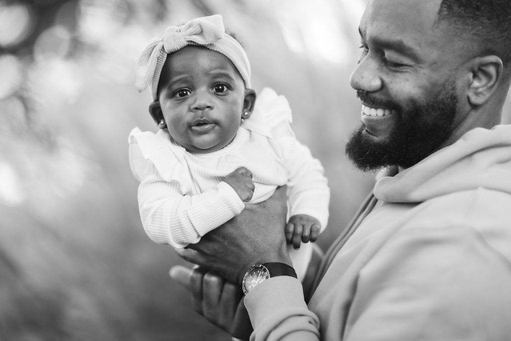 A black-and-white portrait shows a man joyfully holding a baby, who wears a charming headband with a bow. Both are smiling, capturing a heartfelt family moment.