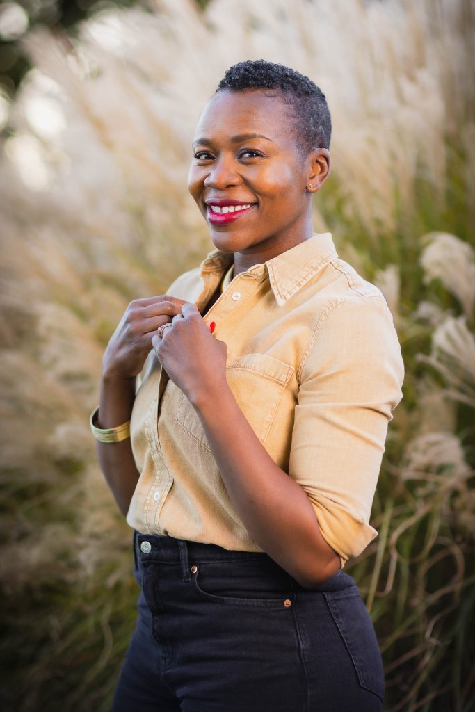 A person with a short haircut smiles while standing outside in front of tall grass, wearing a beige button-up shirt and dark high-waisted pants, creating a warm portrait that exudes a sense of family connection.