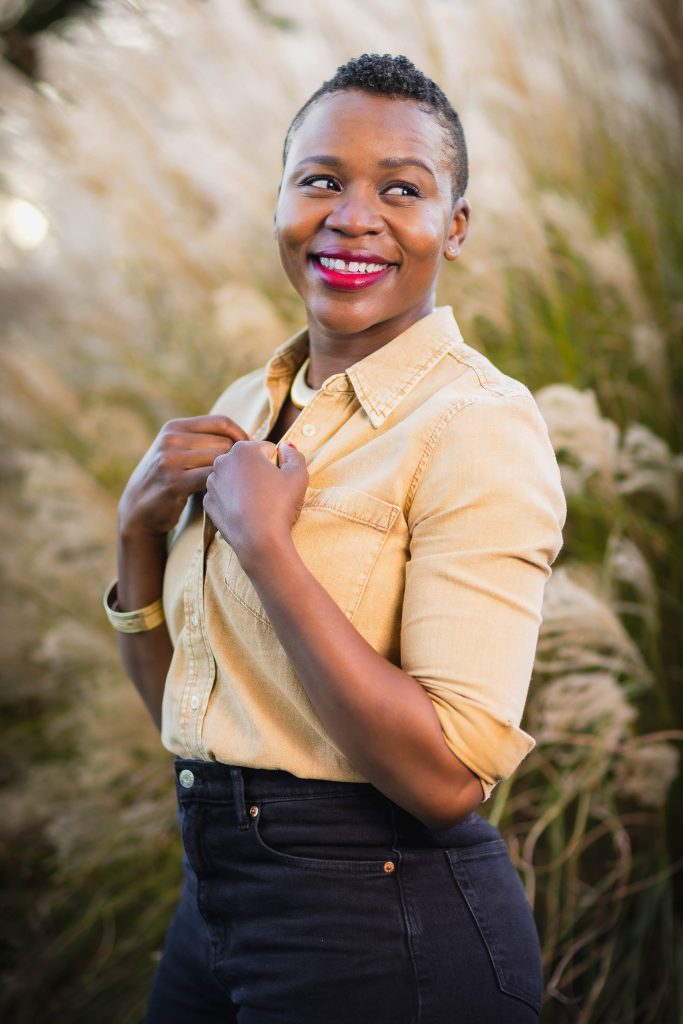 A woman in a yellow button-down shirt and dark pants smiles while standing in front of tall grass, her hands touching her collar, creating a perfect family portrait.